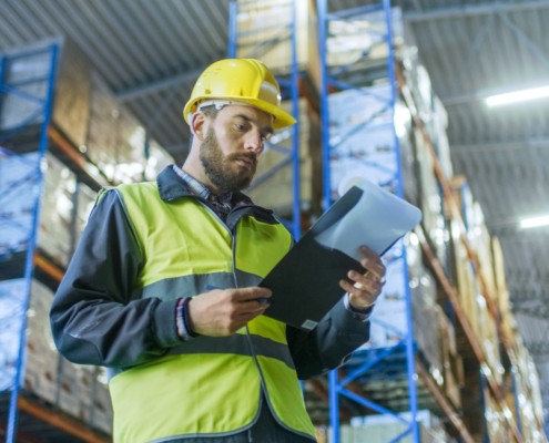 Overseer Wearing Hard Hat with Clipboard Fills in Forms in a Warehouse